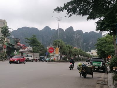 This image captures a street scene in Ha Tu Ward, Vietnam, under an overcast sky, with dramatic karstic limestone mountains forming a prominent backdrop. The light is soft and diffused, suggesting a cloudy day, possibly morning or late afternoon.
In the foreground, a paved road stretches across the frame, indicating an active street or intersection. Several vehicles are present: a red crossover SUV (possibly a Hyundai Kona) is visible on the left, appearing to turn; further down, a white truck and a blue truck are parked or moving slowly. Two motorcycles are also seen on the road, one in the mid-ground and another further back. On the right side of the road, a dark green or black pickup truck is parked by the sidewalk. A person, wearing a conical hat and gloves, is sweeping the sidewalk next to this truck. Nearby, a two-wheeled cart laden with various items, possibly refuse or recyclables, is being pushed by an unseen person or is stationary near the road.
The street is lined with buildings, primarily low-rise structures with visible rooftops and some storefronts. Palm trees are planted along the median or side of the road, and other leafy trees provide shade. Distinctive streetlights with circular, ornate designs are visible in the mid-ground, extending down the street.
Notable street signs include a circular red "do not enter" sign on the median and a yellow sign with "5G Mobifone" partially visible. The pickup truck prominently displays Vietnamese text: "DỊCH VỤ (SERVIE)" and "THAY DẦU", which translates to "Service" and "Oil Change". The overall scene conveys the bustling yet calm atmosphere of everyday life in a Vietnamese town, set against a striking natural landscape.