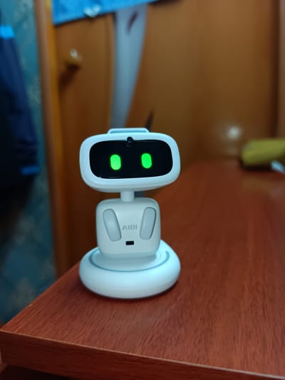 A white robot toy, branded "AIBI," is positioned centrally on a dark wooden table. The robot has a rectangular black screen displaying two green, striped oval shapes, resembling eyes, and a small camera lens above them. Below the screen, the robot has a white neck and torso with two oval shapes on either side, possibly representing arms or speakers, and a small dark rectangle in the center of the torso. The robot stands on a circular white base. The background is blurred, showing a wooden door and a blue textile hanging to the left. The scene is indoors, likely in a domestic setting, and no specific location in Naurovo, Russia can be identified from the visual information provided.