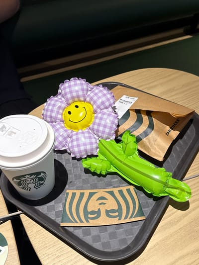 A high-angle shot depicts a black plastic serving tray on a light-colored, possibly wooden, table. On the tray, there is a white paper coffee cup with the Starbucks logo visible. Next to the cup is a purple and white checkered flower-shaped balloon with a yellow smiley face in the center. A partially visible brown paper Starbucks bag is also present, along with a green inflatable toy resembling a crocodile or alligator. A small white paper receipt is visible near the balloon. The background is partially obscured, revealing dark green wall panels and a hint of a person wearing dark clothing. The setting appears to be indoors, possibly a Starbucks store. The image was taken in Hongkou District, China.