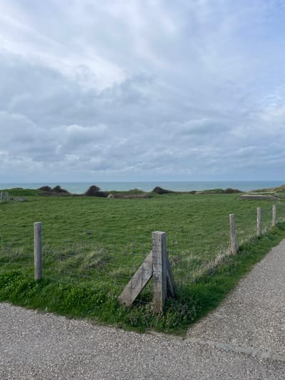 The scene depicts a coastal landscape in Audinghen, France, under an overcast sky dominated by light gray, striated clouds. A vast, verdant green grassy field occupies the mid-ground, extending towards a turbulent, teal-green sea visible along the horizon. Along the coastline, within the green expanse, several low-lying, subtly integrated grey concrete structures, consistent with historical fortifications, are present. In the foreground, a wire mesh fence defines the boundary between the field and a light grey gravel pathway. A prominent corner fence post assembly in the lower center comprises two vertical, weathered wooden posts secured by a diagonal wooden brace, forming an A-frame support structure. Additional wooden fence posts extend to the left and right, holding the wire mesh. The gravel pathway runs along the bottom and right edge of the frame, featuring a subtle, darker grey cobblestone-like border along its inner edge. Sparse green foliage and small flowering plants with purple hues are visible at the base of the fence and along the pathway's perimeter. No individuals or active subjects are present.