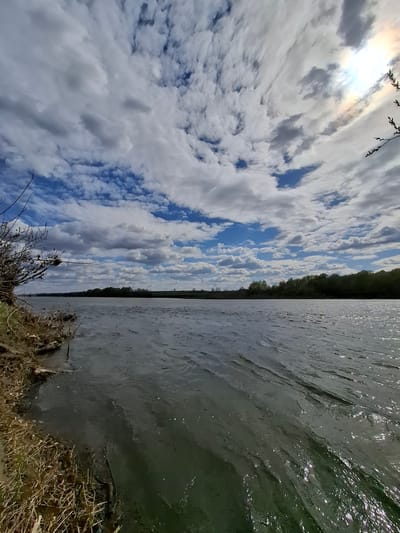 The image depicts a landscape, likely in a rural or natural setting near Дерезовка, Russia. The left half of the image shows a sky filled with white and grey clouds, with patches of blue sky visible in between. The sun is obscured by clouds, but a bright area indicates its location. Some branches are visible in the top left corner of the image. The right half depicts a body of water, possibly a lake or river, with ripples on the surface. A dark strip of trees is seen in the middle of the frame, separating the sky and water. A grassy bank is in the bottom right corner of the image. No people or animals are visible.