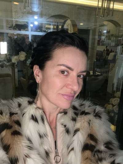 The image displays a head-and-shoulders selfie of a woman, aged approximately 40-50 years, situated indoors within the Smederevska Palanka Municipality, Serbia. The woman has dark hair styled away from her face, prominent brown eyes, and light skin with visible makeup, including pink lipstick. She is adorned with large silver hoop earrings. Her attire consists of a faux fur coat, predominantly white and cream with distinct mottled dark brown or black patches. Beneath the fur, a light pink or beige garment with a silver zipper and a circular pull tab is visible. The background, observed through reflection in a glass or mirrored surface, reveals an interior commercial space, likely a salon or beauty establishment. Visible reflections include shelves stocked with various product bottles, a partially legible sign featuring stylized text, an arrangement of light-colored artificial flowers, and modern interior lighting, including a vertical illuminated panel and a metallic chandelier-like fixture.
