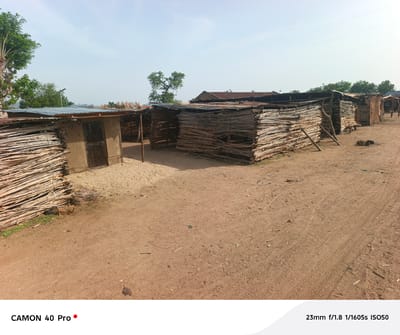 The image depicts a dirt road in Langtang, Nigeria, lined with several small, rudimentary structures. These structures appear to be made from woven branches or sticks, with some having corrugated metal roofs. One structure has a small door. The ground is reddish-brown dirt, and there are tire tracks visible in the dirt road. The sky is overcast. In the background, some trees and possibly hills are visible. There are no people visible in the immediate foreground, but a few figures can be faintly seen in the distance near the last structures. The scene appears to be a rural or underdeveloped area.