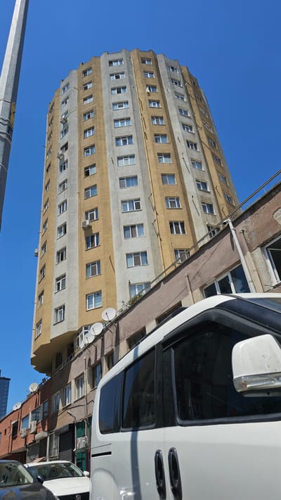 The image shows a tall, multi-story building with alternating bands of yellow and grey concrete. The building has many windows, some of which are open. There are also several satellite dishes mounted on the exterior of the building. The sky is clear and blue, suggesting a sunny day.

In the foreground, a white vehicle, likely a van or bus, is partially visible, obscuring some of the building. The vehicle has tinted windows and a side mirror. The perspective of the image is tilted, making it appear as if the building is leaning.

The scene appears to be an urban setting, with other buildings visible in the background, some with red facades. The presence of satellite dishes suggests a residential or commercial area. No people are visible in the image. The time of day appears to be daytime, given the bright sunlight and clear sky. The location is identified as Ataşehir, Turkey.