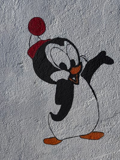 A cartoon penguin character is depicted in a mural painted on a textured, light gray stucco wall in Tomé, Chile. The penguin features a black head, back, and wings, contrasted with a white belly and face. It has large, wide-set eyes with black pupils and an orange beak. A red beanie with a prominent red pom-pom adorns its head. The penguin is shown in an active pose, with its left wing bent and raised towards its face, while its right wing extends outwards. Two orange feet are visible at the base. The outlines of the character are in black.