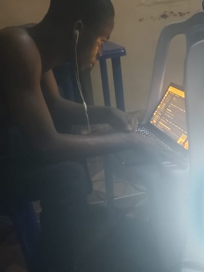 A man sits in a chair wearing earphones and using a laptop computer. The location is unclear.