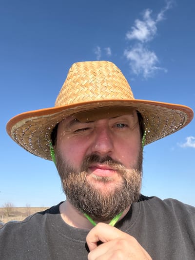 An adult male is depicted outdoors in Зеленогорское сельское поселение, Ukraine. He has a full beard and is wearing a woven straw hat with an orange-trimmed brim and a dark, short-sleeved t-shirt. He is winking his right eye while his left eye is open. His right hand grasps a bright green string, likely a chin strap, near his neck. The scene is illuminated by strong sunlight, casting shadows from the hat onto his face. The background consists primarily of a clear blue sky with minimal scattered white clouds, and a distant, blurred horizon line with faint vegetation is visible to the left.