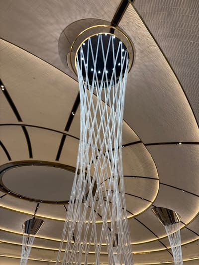 In Winchester, United States of America, the scene is an interior architectural space captured from a low-angle perspective, directed upwards towards a complex, multi-layered ceiling. A prominent, elongated light fixture descends from a central circular aperture in the ceiling. This primary fixture is composed of numerous translucent, tubular elements arranged in a crisscrossing, lattice-like pattern, forming a tapering cylindrical or conical shape. The upper origin point of this fixture is a dark, recessed circular opening, bordered by a golden-colored ring, which contains multiple bright white point lights directed downwards into the translucent structure. The surrounding ceiling is characterized by curved and segmented panels with a textured, light-toned surface, demarcated by dark, thin architectural lines. Warm-toned recessed spot lights are integrated into these ceiling panels, providing ambient illumination. In the lower left and right areas of the frame, two additional, smaller inverted-conical light fixtures are partially visible, also featuring translucent elements, extending from similar circular ceiling cutouts. No people or specific actions are observed. The environment suggests a contemporary, high-ceilinged public interior space.