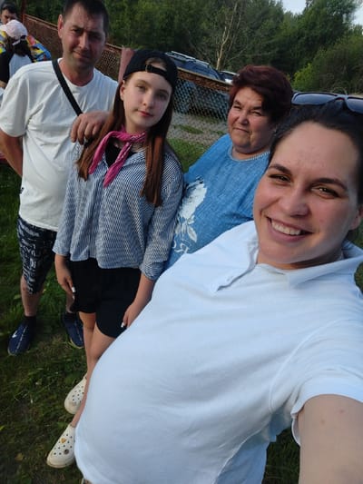 In an outdoor scene, five individuals are captured in a casual, group selfie. The location is identified as Detskii Dom (Bashenino), Russia. In the foreground, a woman in a white polo shirt smiles directly at the camera, her face and upper body filling the right side of the frame. Behind her, a woman with short, reddish-brown hair wears a blue top. To her left, a young girl with a black baseball cap and a pink scarf around her neck is visible. She wears a striped long-sleeved shirt and black shorts. The leftmost figure is a man wearing a white t-shirt and patterned shorts, with a black strap across his chest. Partially visible behind him are other individuals. The group is positioned on grassy terrain, with a fence and parked vehicles in the background, suggesting an outdoor gathering or event. The lighting indicates it is daytime.