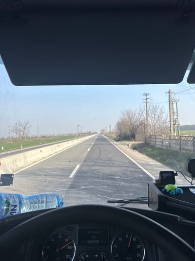 The image captures a forward-facing view from inside a vehicle, likely a truck, in Călugăreni, Romania. The foreground features the vehicle's dashboard, including the top of a black steering wheel and a speedometer indicating approximately 90 km/h. Two plastic water bottles with blue caps, bearing partial "NU" labels, rest on the dashboard. To the right, a black dashcam is mounted, next to a small blue and green toy turtle. The windshield displays dirt smudges and several crack lines.

Beyond the windshield, a two-lane asphalt road extends straight into the distance under a clear, light blue sky. The road has a solid white line marking the right shoulder and a broken white line segment visible in the center of the left lane. On the immediate left side of the road, a series of light-colored concrete barriers with red markings separates the road from a grassy median and a field containing sparse, bare trees. In the farther distance on the left, faint structures are visible.

On the right side of the road, a dirt shoulder is bordered by a concrete fence. Beyond the fence, bare trees and bushes line the road, interspersed with multiple utility poles equipped with transformers and wires. A dirt path and a dark metal gate are visible behind the fence. The overall environment suggests a rural or semi-rural setting during late winter or early spring. No people are visible in the scene.