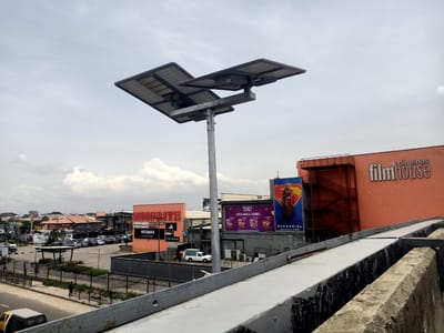 The photograph shows an outdoor scene in Eputu Town, Nigeria. A modern solar-powered street light stands prominently in the foreground, with two rectangular solar panels angled towards the sky. In the background, there are several buildings and commercial establishments. "SHOPRITE", and "filmhouse" signs are visible, as well as an advertisement for the "SUPERGIRL" movie. A parking lot with parked cars is on the left, and a few pedestrians can be seen. The sky is overcast. The image is taken from a slightly elevated position, likely from a bridge or overpass, as suggested by the concrete barrier in the foreground.