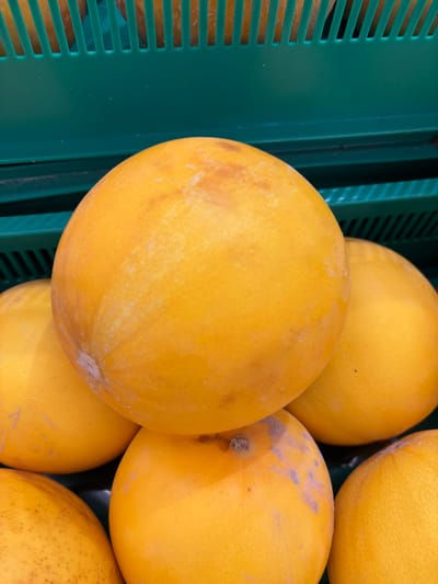 A close-up, overhead view of a pile of bright yellow-orange citrus fruits, likely grapefruits, is presented. The fruits are rounded and have slightly textured skins, with some exhibiting minor blemishes or darker spots. They are stacked in a green plastic bin with a slatted design visible in the background and at the bottom of the frame. The lighting appears to be artificial, illuminating the fruits from above. The image is a still photograph with no visible people or actions. The scene is likely from a market or grocery store in Kuzmolovsky, Russia, given the context provided, though no specific landmarks confirm this location.