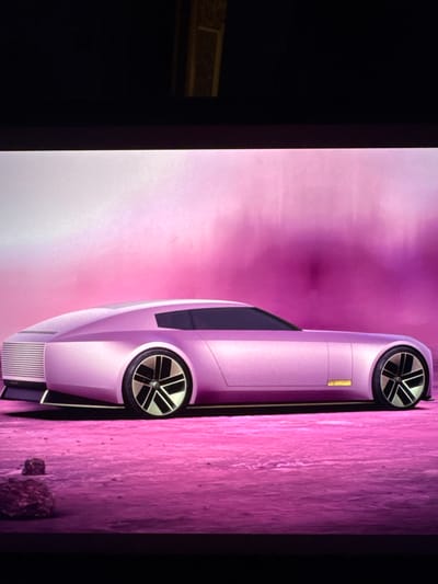 The image is a digital rendering of a futuristic concept car, presented in a profile view. The car is a light pink or lavender color, with a sleek, elongated silhouette. It features a distinctive front grille design, dark tinted windows, and large, multi-spoke wheels with a black and silver finish. The car is positioned on a pink, textured surface that resembles a desert or barren alien landscape, with a few dark-colored rocks scattered in the foreground. The background is a gradient of soft pink and white, suggesting a sky or atmospheric effect. There is no indication of the city of Sant'Angelo di Piove di Sacco, Italy, in this image.