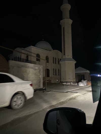 In Miatli, Russia, a white sedan is captured in motion at night, its rear section visible in the foreground, suggesting a forward-moving perspective from within another vehicle. The road appears wet or reflective, illuminated by artificial light sources. Adjacent to the road, a light-colored mosque stands prominently, featuring a large dome and a tall, slender minaret. The mosque is illuminated, highlighting its architectural details and the surrounding construction materials, including sacks of what appear to be cement or sand and stacks of bricks. The environment is dark, indicating nighttime, with the sky a deep black.