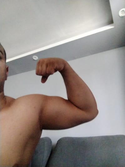 The image shows a close-up of a person's flexed bicep and forearm, with their fist clenched. The person is shirtless, and their skin tone is light brown. The background consists of a white wall and a gray ceiling with recessed lighting. A portion of a gray sofa or chair is visible in the lower right corner. The image is taken from a low angle, looking upwards. There is no information in the image to suggest a location in Kalitinovo, Bulgaria.