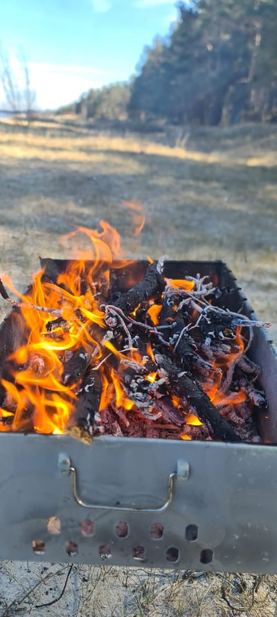 The image shows a metal grill filled with burning wood and branches. The flames are orange and yellow, and the wood is black and charred. The grill is sitting on the ground, with grass and trees visible in the background. The location appears to be outdoors, possibly in Zīvartiņš, Latvia. The image primarily captures a fire burning in a portable grill, in an outdoor setting.