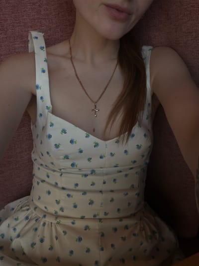 A person is wearing a light-colored dress with a floral pattern of small blue flowers. The dress has thin straps and a sweetheart neckline. A thin gold chain necklace with a cross pendant is visible around the person's neck. The person's hair is light brown and appears to be pulled back. The background is a textured reddish-brown surface, possibly upholstery. The image is cropped to focus on the upper body of the person. There is no indication of the location in Gorzow, Poland.