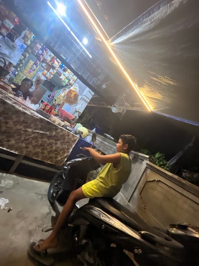 In Mendoyo, Indonesia, a young boy, appearing to be of Indonesian ethnicity, sits on a black motorcycle. He is wearing a bright yellow tank top and yellow shorts. His left foot is resting on the ground, wearing a sandal. The motorcycle is parked in front of a roadside stall or shop. The stall is covered by a dark tarp or awning illuminated by bright yellow LED lights, which cast a strong glare. Behind the boy and to his left, a person with glasses, possibly the shopkeeper, is visible behind a counter laden with various goods, including snacks and packaged items. The background shows a wall and some greenery. The lighting suggests it is evening or night.