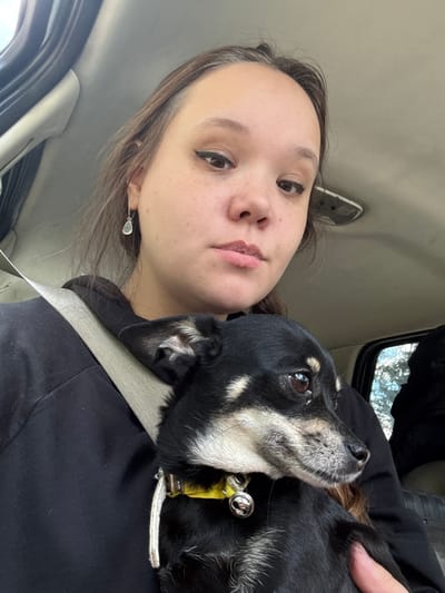 This media file features a close-up selfie of a young person and a small black and tan dog in a car. The person is on the left side of the frame, looking towards the camera with a neutral expression. They have light brown hair styled loosely, and appear to be wearing subtle makeup, including eyeliner. A delicate silver earring with a light blue stone is visible. The dog is on the right, nestled against the person's chest. The dog has dark eyes, a black coat with tan markings around its face and chest, and is wearing a yellow collar with a silver bell. The dog is looking upwards, away from the camera, with a calm demeanor.
The setting appears to be the interior of a car, suggested by the visible car seatbelt, the texture of the car's interior on the left, and the window with a hint of trees and sky visible in the upper right corner, suggesting it is daytime. The overall lighting suggests it is not direct sunlight, possibly overcast or an indoor setting with natural light. The image has a personal, candid feel, capturing a moment between the person and their pet. There is no visible text in the image.