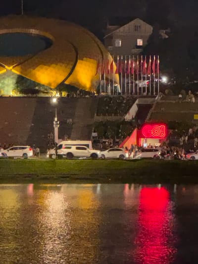 The image captures a nighttime scene in Xuan Huong Lake, Da Lat, Vietnam, showcasing a vibrant display of lights and activity. In the background, a large, futuristic, golden-domed structure is illuminated, casting a warm glow. Adjacent to it, a row of red and yellow flags flutters against the dark sky, hinting at a festive occasion. Further down, a building with illuminated windows stands on a hillside, nestled amongst trees.
In the midground, a street is lined with several white vehicles, including SUVs and vans, suggesting a gathering or event. Pedestrians are visible, some milling about and others possibly entering or exiting a building. A prominent neon sign in bright red, spelling out "GO!", is affixed to a building facade, further indicating a lively atmosphere. The reflection of the lights and the surrounding environment shimmers on the surface of Xuan Huong Lake in the foreground, creating a dynamic and colorful tableau. The overall impression is one of a bustling urban nightscape with elements of modern architecture and public celebration.