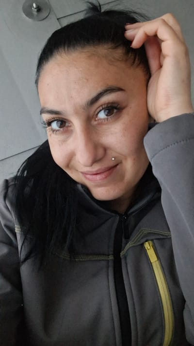 A close-up, medium shot shows a woman from the chest up, looking at the camera. She has dark hair pulled back, and is wearing a gray jacket with a yellow zipper. She is resting her right hand on her head and smiling softly. She has a small stud piercing below her lower lip on the left side of her face. The background appears to be an indoor setting with a plain wall and what looks like a light fixture on the ceiling. The image was taken in Račice, Czechia.