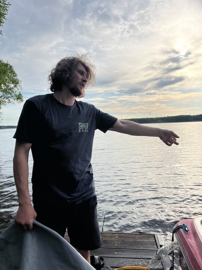 A man with shoulder-length curly hair and a beard is standing on a wooden dock next to a body of water. He is wearing a dark t-shirt and shorts. His right arm is raised towards the sky. A boat is visible on the right side of the frame, partially on the dock. The sky is cloudy. There is green foliage visible on the left side of the frame, suggesting trees or shrubs. The image appears to be taken near Пятница, Russia.