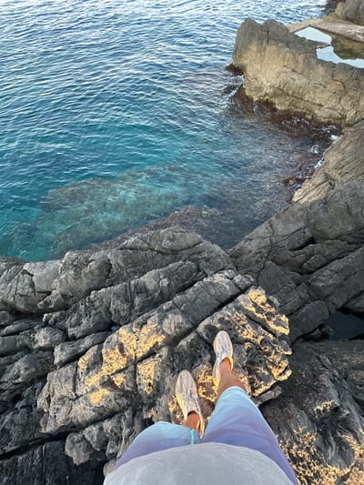 The image shows a first-person perspective looking down at a person's legs and feet standing on rugged, rocky terrain. The person is wearing patterned slip-on shoes and light blue shorts over a gray garment. The rocks are dark gray, with areas illuminated by sunlight, creating a contrast of light and shadow.

Beyond the rocks, the scene transitions to a body of clear, turquoise water. The water appears shallow in some areas, revealing the rocky seabed beneath, and deeper and darker in others. The surface of the water is rippled, indicating gentle movement. In the upper right portion of the image, a rocky outcrop extends into the water, with some flat, man-made sections that appear to form shallow pools or platforms, possibly part of a coastal structure. The overall environment suggests a natural coastal setting, likely in or near Višnjevo, Montenegro, given the characteristic rocky coastline and the clear waters of the Adriatic Sea.