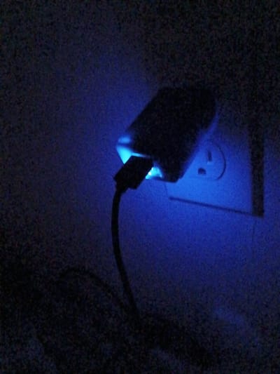 A low-light photograph shows a white electrical outlet with a USB charger plugged into it. A USB cable is attached to the charger. The light emitting from the charging device casts a blue hue on the scene. The location is not discernible from the image.