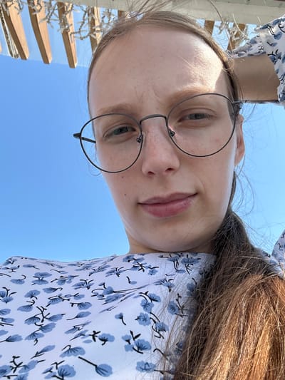 A close-up, low-angle selfie of a woman with long brown hair, wearing round glasses and a white dress with a blue floral pattern. She is outdoors with a clear blue sky in the background and what appears to be a wooden structure with lights overhead. The woman's right hand is resting on the back of her head. The setting is a sunny day, possibly in Металлплощадка, Russia.