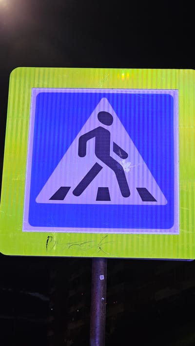 A pedestrian crossing sign is mounted on a metal pole, set against a dark, blurred background suggesting it is nighttime in an urban environment, possibly Yekaterinburg, Russia. The sign itself is triangular with a blue interior bordered by a thicker white outline and a luminous green border with vertical stripes. Inside the blue triangle, a dark silhouette of a person is depicted walking across a crosswalk, indicated by parallel dark rectangular shapes. The sign shows some minor scuff marks and what appears to be graffiti in black ink on the lower part of the green border. A small white square sticker is affixed to the metal pole supporting the sign. The lighting suggests artificial illumination.