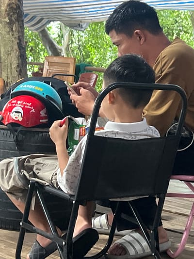 Two individuals, a man and a boy, are seated in chairs outdoors, partially shaded by a striped tarp. The man, in the background, wears a brown shirt and is looking to his left. The boy, in the foreground and facing away from the camera, wears a patterned white shirt and shorts. He is holding a red and white object in his right hand. In front of them, on what appears to be stacked tires, are two helmets, one blue with "XANHSM" visible and another red one with a cartoon character and the Honda logo. The environment suggests a casual outdoor setting, possibly a roadside rest area or a home's outdoor space. The ground is a wooden deck. The overall scene is unposed and appears to capture a candid moment. The specific location within Tam Dương Bắc, Vietnam is not ascertainable from the visual information provided in the image.