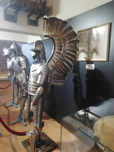 The image displays an indoor museum exhibit within the Mir Castle Complex, located in Мірскі сельскі Савет, Belarus. Three full suits of historical armor are presented on display. In the immediate foreground, a Winged Hussar armor stands prominently, featuring a large, elaborately feathered wing attached to its back and detailed decorative elements on its breastplate. This armor is supported by a dark wooden base. Two additional suits of plate armor are positioned behind and slightly to the left of the Winged Hussar armor, mounted on metal stands. A red velvet rope, supported by chrome stanchions, defines the exhibit boundary on a light-brown tiled floor. The background to the right features a dark gray wall, upon which a gold-framed historical map and a small white information plaque are mounted. A brick wall is visible further left in the background. Overhead, industrial-style architectural elements are suspended from a light-colored ceiling. The lower right foreground reveals a corner of a clear glass display case, partially containing a light-colored, rounded object. Shadows from the exhibits, notably a distinct feather pattern from the Winged Hussar's wing, are cast upon the dark wall.