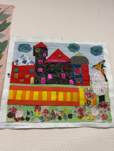 This image presents a textile art piece, identified as a collaborative work from the "Young Ecologist" association in Novosergievka, Russia, led by E.S. Chepik. The artwork is displayed against a light, textured white background, with a portion of another textile piece visible on the left.
The main subject is a rectangular appliqué or collage depicting a fantastical cityscape. The upper section features a light blue fabric sky with two irregular dark teal fabric clouds. Three distinct buildings comprise the city:
1. **Leftmost building**: Constructed from red fabric, with a white fabric section patterned with blue flowers. Its roof is red, and it features a single teal button and several gold metallic horizontal strips. A blue pushpin secures a portion of this building to the backing.
2. **Central building**: Taller than the others, it is made of black fabric with a gold leaf pattern. It has a pointed red roof adorned with a small multicolored bead. Multiple buttons (brown, green, red) and horizontal pink metallic strips are applied as decorative elements.
3. **Rightmost building**: Features a red fabric base, topped by a dome-shaped section of grey and black houndstooth fabric. Below the dome, a blue fabric section with vertical blue metallic strips and a brown button represents a window. The building is topped with a pointed red fabric roof.
Below the buildings, a horizontal strip of green fabric represents the ground. A prominent fence or wall, composed of alternating yellow and orange vertical strips bordered by solid orange horizontal strips, spans the mid-ground. To the right, a stylized tree with a dark brown trunk and a dense white floral canopy is visible. A yellow bird with black markings is perched near the top of this tree. The foreground along the bottom edge of the green fabric is adorned with numerous cut-out paper or fabric floral shapes in various colors and sizes, alongside some small butterfly cutouts.
The artwork itself has a rough, white-painted or glued border and is held in place by visible pushpins, one of which is at the upper right corner.