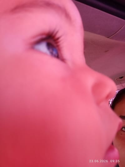 The image is a close-up, low-angle shot of a young child's face, predominantly in pink hues, suggesting an artificial light source or filter. The child's left eye, with its blue iris and long eyelashes, is in focus and looking towards the right of the frame. A portion of a car's interior, specifically the ceiling, is visible in the upper right quadrant, indicating the setting is likely within a vehicle. In the lower right corner, another person's face, an adult or older child, is partially visible, with their dark hair and right eye discernible. A timestamp "23.06.2026 09:35" is overlaid on the image in the bottom right. The overall scene is intimate and appears to capture a candid moment. There is no indication of the city of Batken City, Kyrgyzstan.
