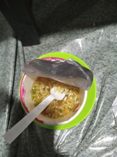 An overhead, slightly angled photograph depicts a partially consumed cup of instant noodles. The cup, predominantly white with pink accents, rests on a bright lime-green plastic saucer or plate. The metallic foil lid of the noodle cup is peeled back towards the top of the frame, exposing the golden-yellow wavy noodles submerged in a light brown broth. Visible within the broth are green specks, likely chopped vegetables or scallions, and a single reddish-brown piece, possibly a slice of sausage or meat, positioned towards the lower right within the cup. A white plastic spork-like utensil is inserted into the noodles, with its handle extending towards the lower-left corner of the image. The immediate surface beneath the cup and saucer is a grey, textured material, possibly flooring or linoleum, which is covered by a clear, crinkled plastic sheet, creating reflections and folds. In the upper portion of the background, a dark, out-of-focus rectangular structure, possibly part of furniture or a base, is visible. The specific city of Keudealuerheung, Indonesia, or any geographical location, cannot be identified or confirmed from the visual information present in the image. There are no individuals or discernible actions beyond the implied preparation and consumption of the noodles.