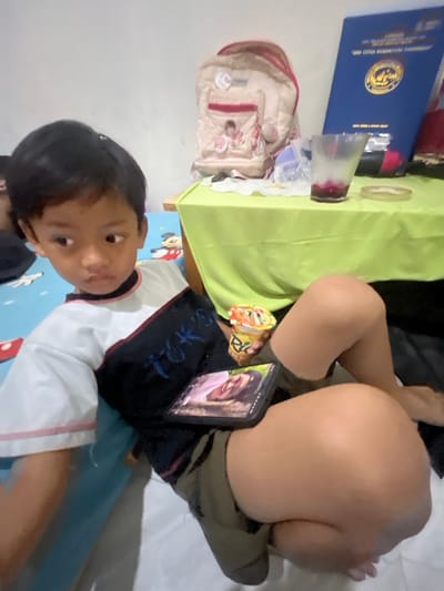 The image shows a young boy sitting on a bed with a phone resting on his lap. He is wearing a t-shirt with a white and dark blue design. A cup of yogurt is next to the phone. In the background, there is a green tablecloth draped over a table, a backpack, and a blue folder. The scene appears to be indoors, likely a bedroom, based on the bed and the wall. The presence of a phone and yogurt suggests a casual, home environment. No specific landmarks in Serdang Kulon, Indonesia, are visible in this image.