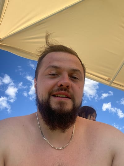 The image is a selfie taken outdoors under a tan umbrella. A man with a beard and wearing a silver chain is the main subject, smiling at the camera. Behind him, a person with dark hair can be seen, partially obscured by the man's shoulder. The background consists of a bright blue sky with scattered white clouds. The location is not specified in the image; it is not possible to determine if it is in Колокша, Russia.