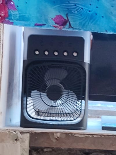 A black and silver portable fan sits on a ledge beneath a blue cloth with purple floral patterns. The fan has a black front grille with a silver circular pattern and four silver buttons on the top. The ledge is made of wood and appears to be part of a window sill or shelf. The background is out of focus, but shows what appears to be a dark surface, possibly a computer monitor, and a blue object that may be a light. The scene appears to be indoors.