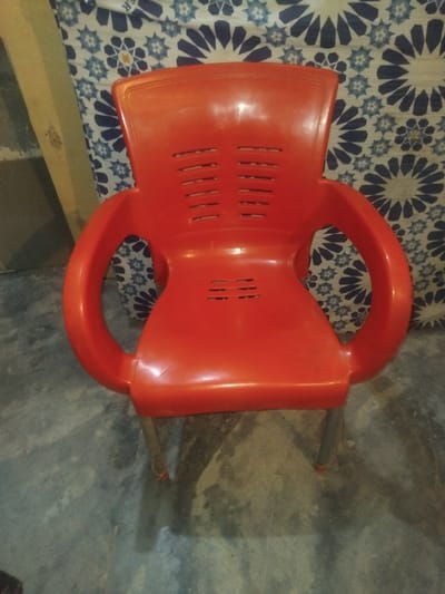 The image shows a bright orange plastic chair placed on a patterned fabric, likely a bedsheet or rug. The fabric features a repeating geometric design of blue and white star-like shapes. The chair has a simple design with a molded seat and back, and armrests that curve downwards. There are slots or vents on the backrest of the chair. The chair is positioned at an angle, casting a shadow on the grey, textured floor. A thin metal rod, possibly a leg of the chair, extends from the side and rests on the floor. The lighting suggests it might be indoors, possibly in a room with natural light from a window or artificial lighting. The setting appears to be simple and perhaps a bit rustic, given the texture of the floor. There is no clear indication of the time of day or weather conditions. No people are visible in the image. The overall impression is a still life composition of a common household object in a domestic setting.