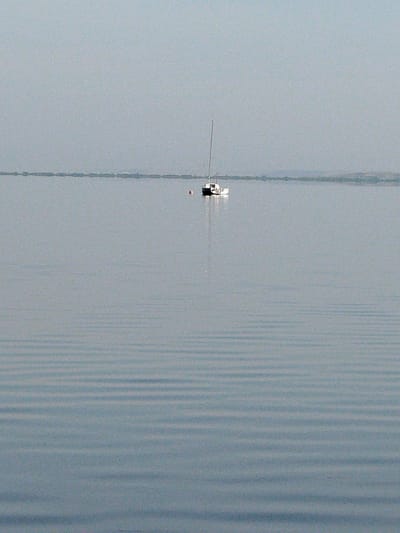 A sailboat with a single mast is moored on a calm, overcast body of water. The water exhibits subtle, horizontal ripples, creating a textured surface. The horizon is low and indistinct, with a faint, hazy outline of land visible in the distance. The sky is a uniform pale gray, devoid of clouds. A small, red buoy is visible near the sailboat. The overall scene conveys a sense of stillness and tranquility. The location is a large, natural body of water, possibly a lake or reservoir, consistent with the geography of Kokshetau, Kazakhstan, which is situated on the shores of Lake Kokshetau.