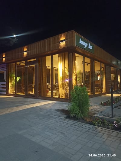 This media file captures an exterior view of a modern, wooden building at night, identified by the sign "lounge bar" in light green lettering. The building features extensive glass windows, offering a view into a warmly lit interior furnished with tables and chairs, suggesting a restaurant or bar setting. Inside, patrons are not clearly visible, but the presence of a bar area with bottles and shelves can be observed. Golden curtains hang within the windows, adding to the inviting ambiance.
Outside, the ground is paved with interlocking bricks, and a paved walkway runs along the front of the building. A small, neatly trimmed evergreen bush stands prominently in the foreground. In the distance, the moon is visible in a dark sky, partially obscured by clouds, and a faint light beam emanates from it. The overall impression is one of a tranquil evening at a well-lit establishment.
The visible text at the bottom right corner of the image reads "24.06.2026 21:40," indicating the date and time the photo was taken. The location context provided is Шұбартүбек, Kazakhstan. The scene evokes a sense of calm and an inviting atmosphere for evening patrons.