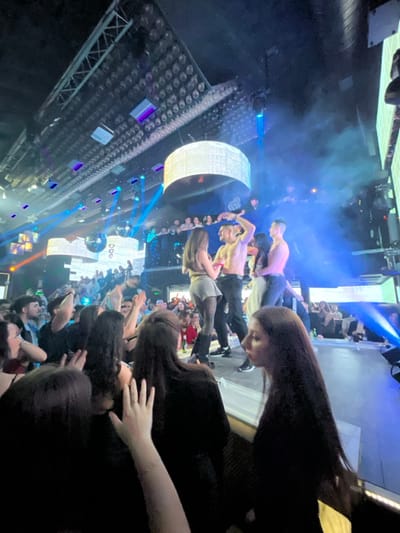 This image depicts an interior scene within a nightclub located in Pamporovo, Bulgaria. The central focus is a raised stage where approximately four to five individuals are actively performing or interacting under dynamic blue lighting and haze. Two shirtless men are visible on stage; one is positioned centrally with his right arm raised, facing a woman wearing light-colored shorts and dark tights. Another shirtless man stands to the right, facing a woman in a light-colored garment.
Above the stage, a large, circular, illuminated fixture hangs from the ceiling, which is equipped with an extensive truss system holding numerous grid-patterned lights and sound equipment. Several disco balls are visible, reflecting light beams. Blue laser beams and spotlights crisscross the venue, contributing to the club atmosphere. Digital screens are visible on the far walls.
A large crowd occupies multiple levels of the venue, observing the stage. Individuals in the immediate foreground, mostly seen from behind or in profile, are part of the audience, with some hands raised, indicating engagement. Spectators are visible both on the main floor in front of the stage and on elevated balconies surrounding the performance area.