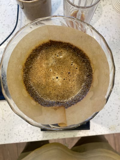 A top-down view reveals a glass pour-over coffee maker with a paper filter. Dark, foamy coffee fills the center, with a ring of saturated grounds around the edge. The filter is wet and clings to the glass. The pour-over device sits on a dark surface, possibly a scale. Adjacent to the coffee maker are blurry images of glass and ceramic vessels, possibly related to the brewing process. The background shows a speckled countertop and a blurry, light-colored object.