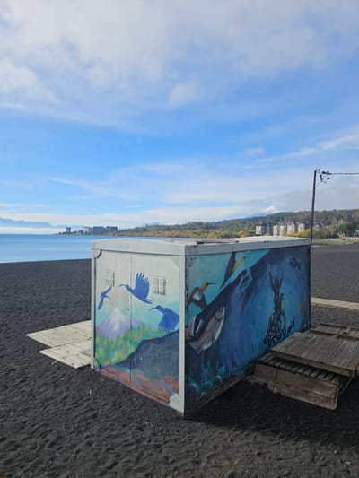 The image depicts an outdoor scene in Villarrica, Chile, likely a beach area. The main subject is a box-shaped structure or utility building with colorful murals on its sides. One mural appears to depict underwater scenes with fish and a diver, while another shows birds in flight. The structure is situated on a dark, sandy surface, presumably a black sand beach, and is partially connected to a weathered wooden platform or walkway. In the background, across a body of water (likely Lake Villarrica), lies a landscape of buildings, trees, and possibly mountains. The sky is bright blue with scattered clouds. A utility pole with wires is visible near the structure. The overall impression is a blend of urban and natural elements, suggesting a location where a city meets the lake.