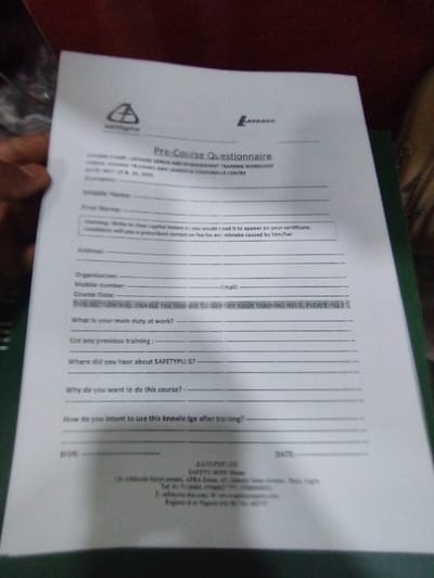 The image shows a pre-course questionnaire form for a "LAGOS MINES AND MANAGEMENT TRAINING WORKSHOP" at the "SAFETYPLUS LEARNING AND CONFIDENCE CENTRE." The form is partially filled out by hand, with the top of the paper being held by an obscured hand. There are blank fields for name, middle name, address, organization, mobile number, and email. The form asks for the individual's main duty at work, any prior training, where they heard about "SAFETYPLI," their reason for taking the course, and how they intend to use the knowledge after training. The form is signed "SAFETYPLUS" with the date field blank. The image does not provide any details about the person filling out the form. The location of Ashaka, Nigeria, with coordinates (latitude: 10.929, longitude: 11.476) does not appear in the image.