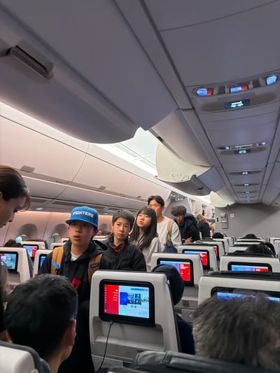The image depicts the interior of a commercial aircraft cabin, specifically an aisle filled with standing passengers, likely during boarding or deplaning, associated with travel to or from New Chitose Airport (CTS), which serves Tomakomai, Japan. The environment is uniformly lit by overhead cabin lights. Overhead bins are closed along both sides of the cabin. Passenger service units are visible along the ceiling, displaying illuminated "fasten seatbelt" and "no smoking" indicators.

In the mid-ground, a group of five individuals stands in the aisle. From left to right: a young male wearing a blue baseball cap with "FIGHTERS" emblazoned on it and a brown backpack, appearing to look forward with his mouth slightly open; a young male with short, spiky dark hair and what appear to be goggles or headphones around his neck; a young female with long dark hair; an adult male with dark hair and a light-colored long-sleeved shirt; and another young male with curly hair wearing a dark jacket. All five are facing generally forward in the aisle. The head and shoulder of a person with dark hair are visible in the immediate foreground on the left.

The backs of several aircraft seats are visible in the foreground and mid-ground, equipped with illuminated in-flight entertainment (IFE) screens. One screen in the center-right shows a menu interface with various content options and a connected charging cable. Further down the aisle, additional passengers are visible, some partially obscured. No direct interactions between individuals are clearly discernible; the primary action is the collective standing in the aisle.