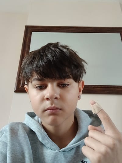 A close-up, rotated portrait captures a young person with short, dark brown hair and dark eyes, looking directly at the camera. They wear a light blue or grey hooded sweatshirt and a small, circular silver earring in their visible left ear. The person's left hand is raised, with the index finger prominently featuring a light-colored band-aid or plaster. Their expression is neutral.
The background consists of a light-colored wall with a wooden-framed mirror positioned to the right of the person's head. The mirror reflects a soft, light-toned area, possibly an opposite wall or empty space. The image appears to be taken indoors under ambient lighting, given the soft illumination and lack of harsh shadows. The overall composition suggests a casual, perhaps self-taken, photograph. There is no visible text in the image. The location context provided is Armagh, United Kingdom, though no specific visual cues for this location are present in the photo.