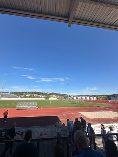 From an elevated vantage point beneath a corrugated metal roof, the image captures a soccer field in Valpaços, Portugal. The field is characterized by green grass, a running track with red lanes, and a sheltered dugout. Adjacent to the field are a series of buildings. Spectators are partially visible behind a metal fence, viewing the activities on the field. Advertisements are affixed to the fence, displaying local businesses and contact information. The backdrop features a clear blue sky dotted with scattered clouds.
