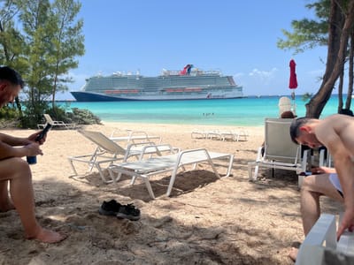 The image shows a sunny day at a beach in the city of Freeport, Bahamas. In the foreground, two men are relaxing on lounge chairs under the shade of trees. The man on the left is looking at his phone and holding a blue can. The man on the right is also looking at his phone. Several white lounge chairs are scattered across the sandy beach. In the background, a large cruise ship, identified by the "Carnival" logo, is docked in the turquoise waters of the ocean. The water is clear and calm, and there are a few people swimming in the distance. The sky is blue with a few scattered clouds. The overall scene depicts a leisurely vacation atmosphere.