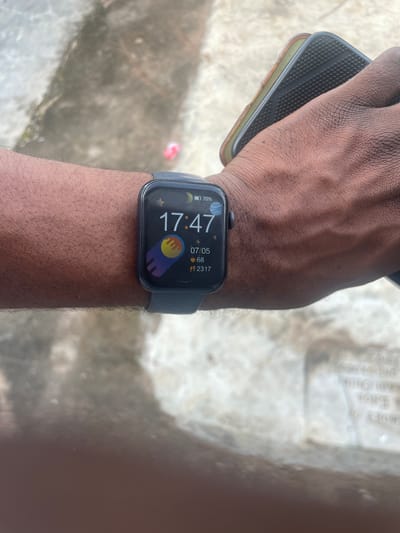 The image shows a person's arm wearing a smartwatch with a dark gray band. The watch face displays the time as 17:47, a battery icon showing 70% charge, the date 07/05, and two stats: 68 and 2317. There is a colorful graphic of a planet and a shooting star on the watch face. The person's hand is holding a smartphone with a textured black case. The background appears to be an outdoor setting with a rough, textured surface, possibly concrete or pavement, and some light-colored stones or debris. There is a small, blurred red object in the background on the ground. The image appears to have been taken in the city of Orodo, Nigeria, given the context provided.